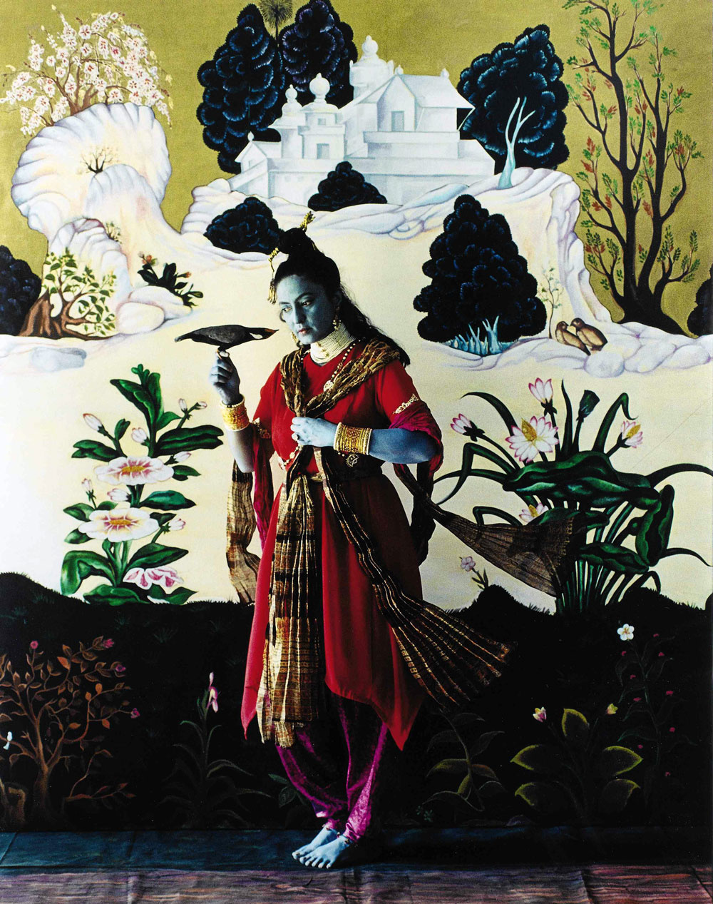 A painting with plants, flowers, and trees leads to a simple white temple with a yellow background. Against this is a photo a strong Indian woman with blue skin dressed in a traditional red sari. She holds a black bird in her right hand, staring at it as the bird stares back.