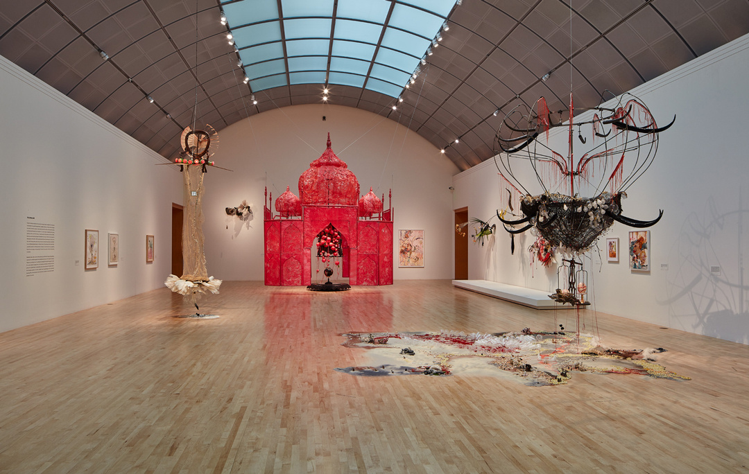 An installation view into a long barrel-vaulted gallery shows small paintings and large-scale sculptures hanging from the ceiling. The hanging works include a floating bright pink temple and a nest-like structure that hovers over a landscape of beach sand on the gallery floor.