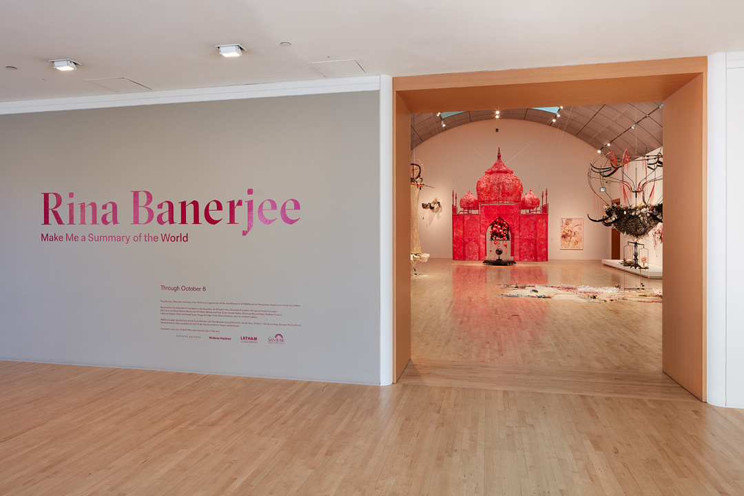 An installation wall shows an entrance to the exhibition "Rina Banerjee: Make Me a Summary of the World" which is written in bright pink letters on a gray wall. Through an adjacent doorway a bright pink 3-dimensional temple is at the back of a barrel-vaulted gallery.