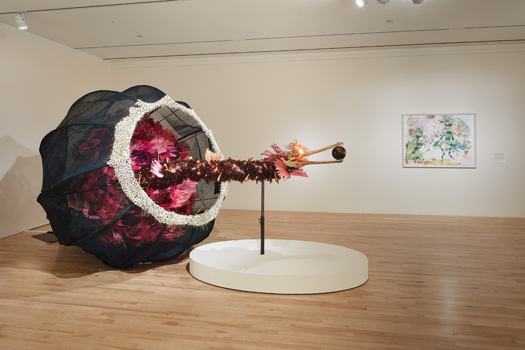 A human-sized black basket filled with large straw flowers sits on its side in the corner of a gallery. Emerging from its center is a long snake-like growth that appears vegetative except for the glowing eyes and tweezer-like beak at its end which holds a globe-like orb.