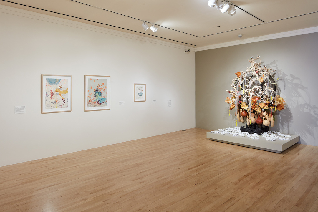 A gallery space shows three small framed paintings on the left wall and a sculptural work on the right. The sculpture resembles a tree with branches, yellow and orange feathery leaves, and wooden gourds hanging downwards. 