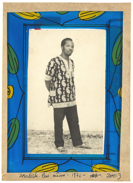 A cream and black photo of a black man holding his relaxed arms behind his back. He looks at the camera, with his body faced to the side, with one leg leaning forward. The photo is surrounded by a blue frame that features green and yellow leaves.