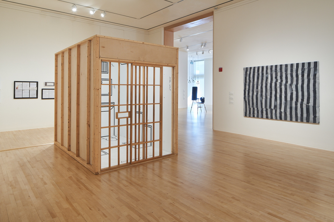 In a gallery is a wooden structure, resembling a tiny room. It has a door of intersecting vertical and horizontal wooden bars. Its inner walls are white with black outlines of obscured items. To the right is a painting of distorted, black and white vertical lines.