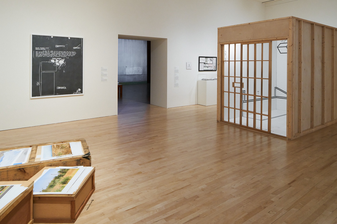 In a gallery is a wooden structure resembling a tiny room. To the left are 3 short wooden crates with prints of horizontal landscapes on top of them. On the walls are 2 framed black and white artworks. Towards the gallery's center, a doorless entryway leads to a shadowy room.