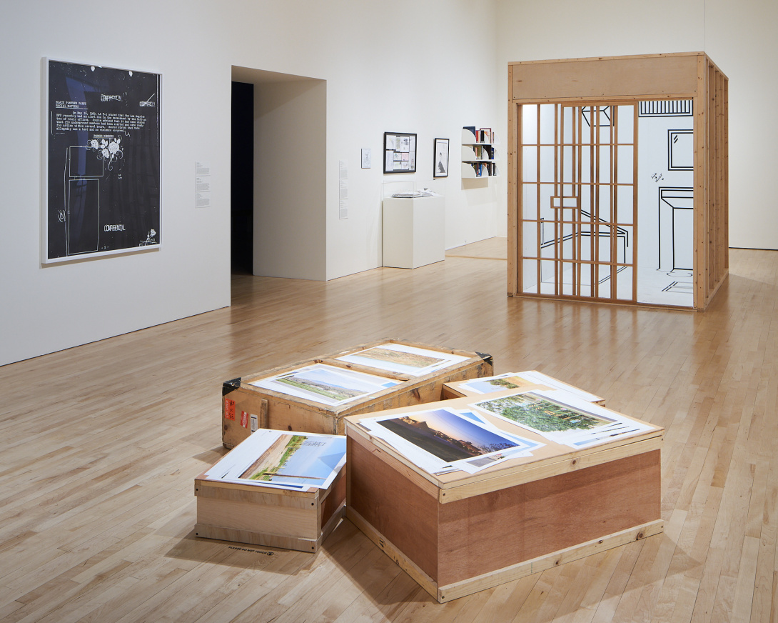 Four short wooden crates have landscape prints resting on them. A wooden structure resembling a tiny room is further back in the gallery. The structure's front has intersecting wooden bars, revealing black outlines of furniture inside. On the walls are framed black and white artworks.