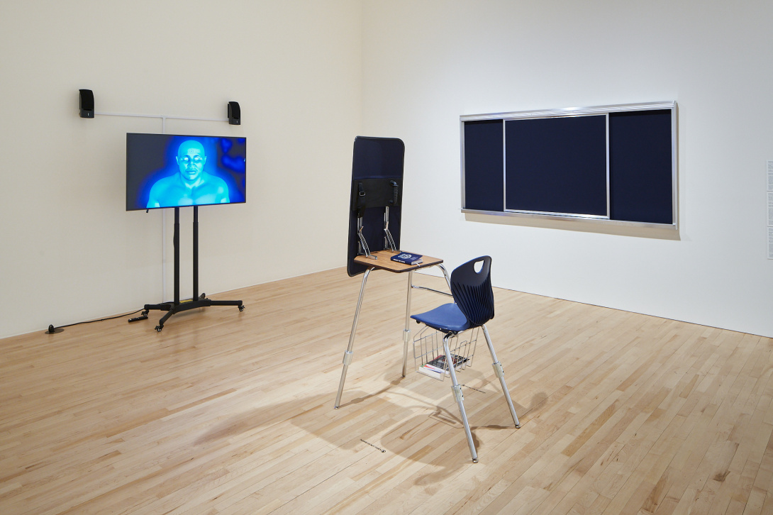 A school desk and chair with an attached riot shield face towards a TV. A glowing, ghostly, blue bust of a figure is seen on the TV, staring with empty eyes towards the desk. A blank black chalkboard is situated on the wall next to the desk,