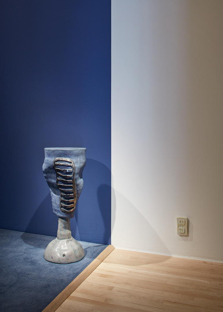 A vertical blue squiggly nondescript sculpture with a silver grate attached to it, sitting on the edge of a blue carpeted gallery with blue walls. On the wall is a electrical outlet ceramic sculpture. 
