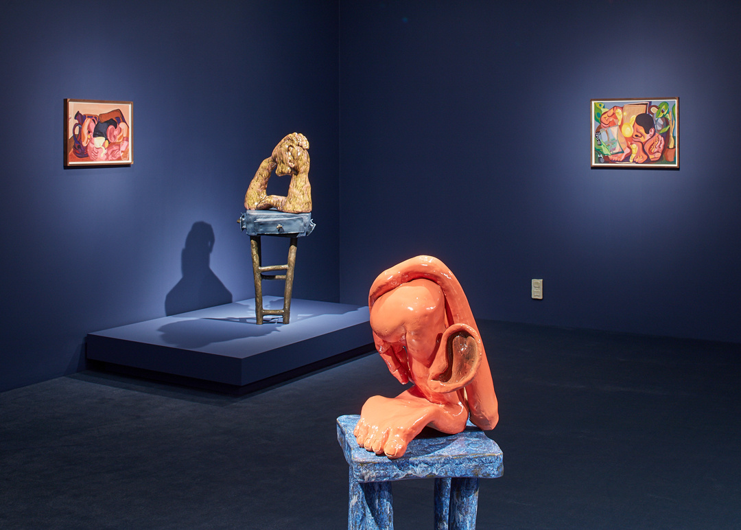 Dark blue painted gallery with two spotlighted ceramic and colorful sculptures on the ground.  Both sculptures are sitting on stools and are very abstract. One appears to have a large foot and an ear. There are two spotlighted paintings on the wall.