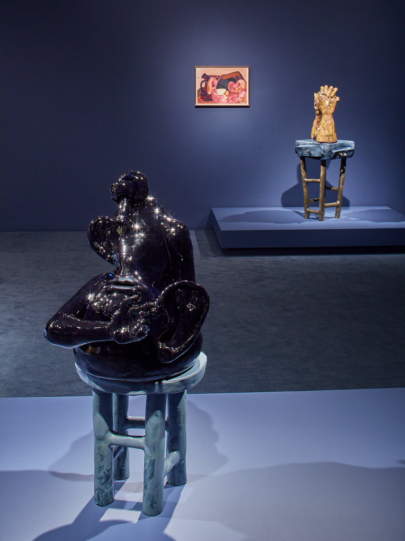 A dark gallery with walls and floor painted dark colors. In the front is an abstract sculpture on a ceramic stool. In the background is a pedestal with another ceramic sculpture on a stool with a painting. There is dramatic lighting on all three pieces.