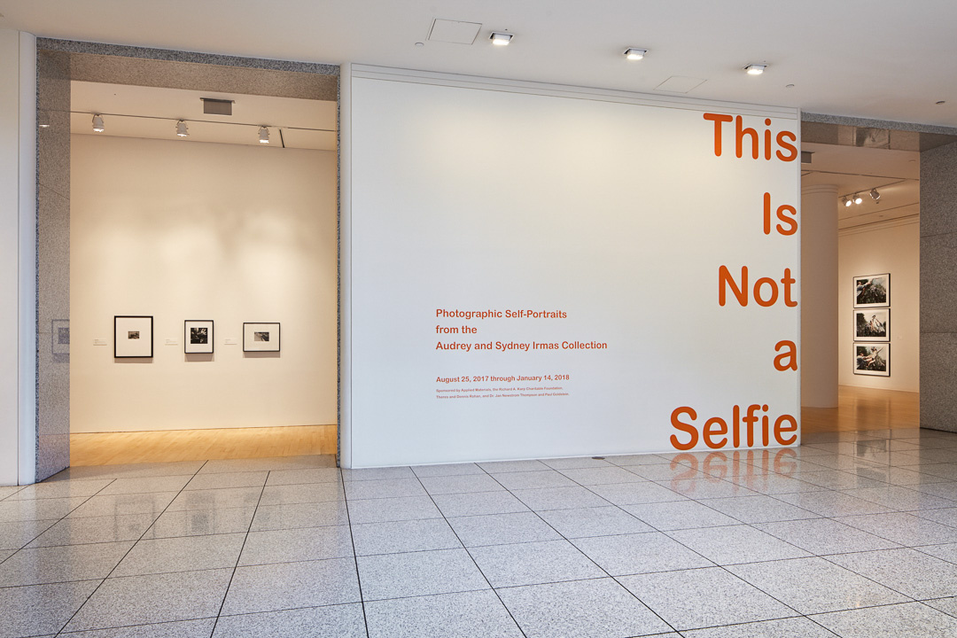 The entryway of a museum gallery. On the wall is the title, This is Not a Selfie, in orange font. On either sides are open spaces, with framed artwork hanging in the background.