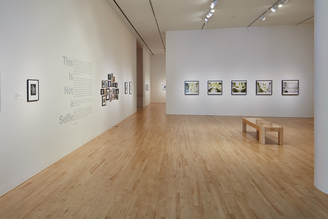 A gallery filled with black and white framed photographs. In the foreground is a wooden bench. To the left another gallery is partially visible, also with art on its walls.  "This is Not a Selfie" is on the wall with many smaller framed photographs grouped together.