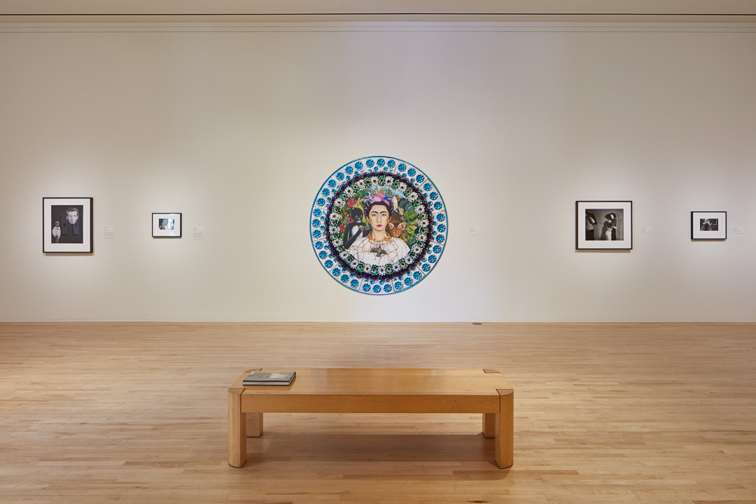 A round painting of a person who looks like Frida Kahlo, but it is not Kahlo's work. A monkey sits on the Kahlo's lookalike's shoulder. On either side of the portrait are much smaller black and white photographs portraits of people.