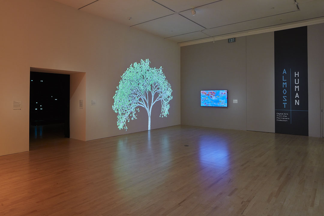 In a gallery is an entry way to another dark gallery with lots of little glowing lights. Next to the entry, a tree is projected onto the wall. A digital screen is installed next to the tree. To the left, text reads “Almost Human” which is applied vertically to the wall.