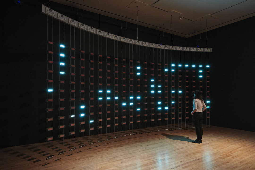 In a dark cavernous room, a single person looks at a curved sculpture, which hangs from the ceiling in a curve against the wall. It is comprised of hundreds of tiny rectangles, designed to hang vertically. Some of them are lit up and emanate an eery turquoise color.