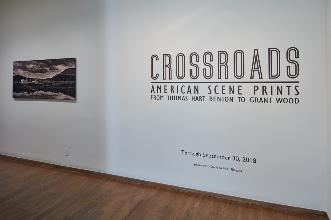 A white wall with a single black and white print of mountains hangs on it.. Also on the wall in vinyl reads "Crossroads American Scene Prints From Thomas Hart Benson to Grant Wood."
