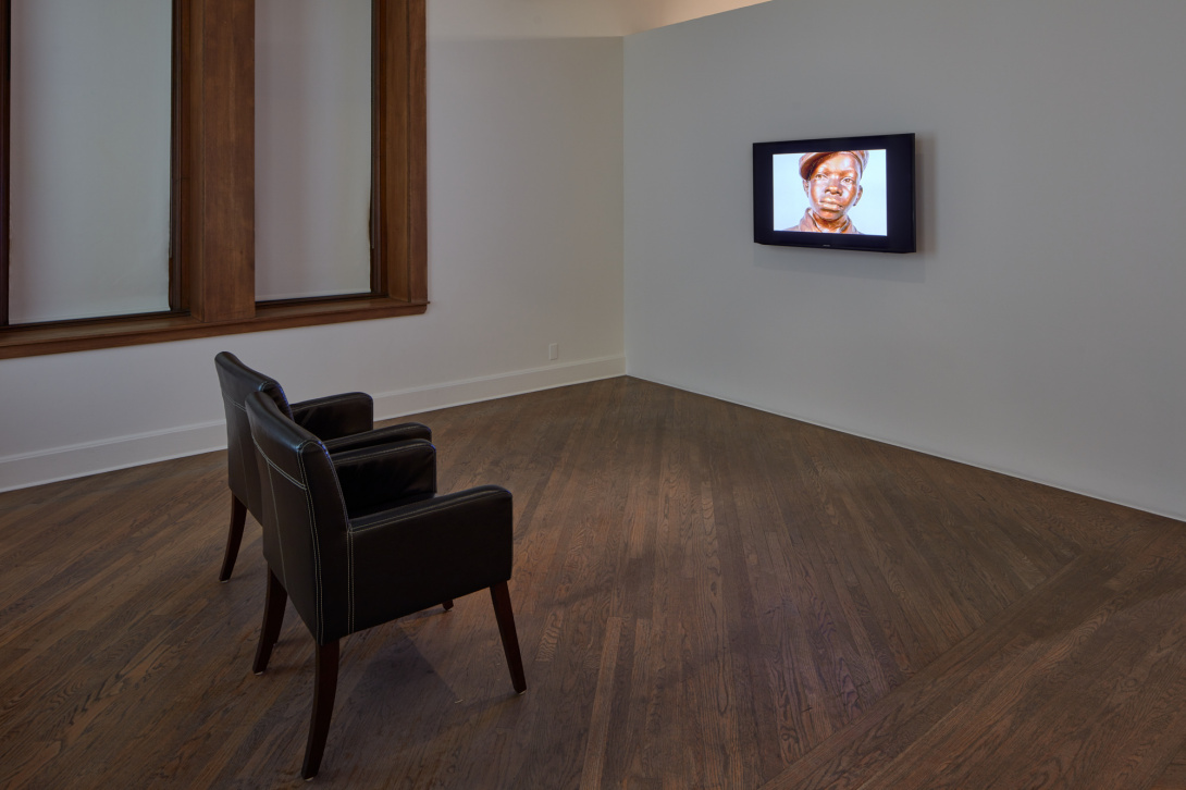 A photograph of two leather chairs facing a single small screen framed with dark borders. It displays a sculpture of a young African male.