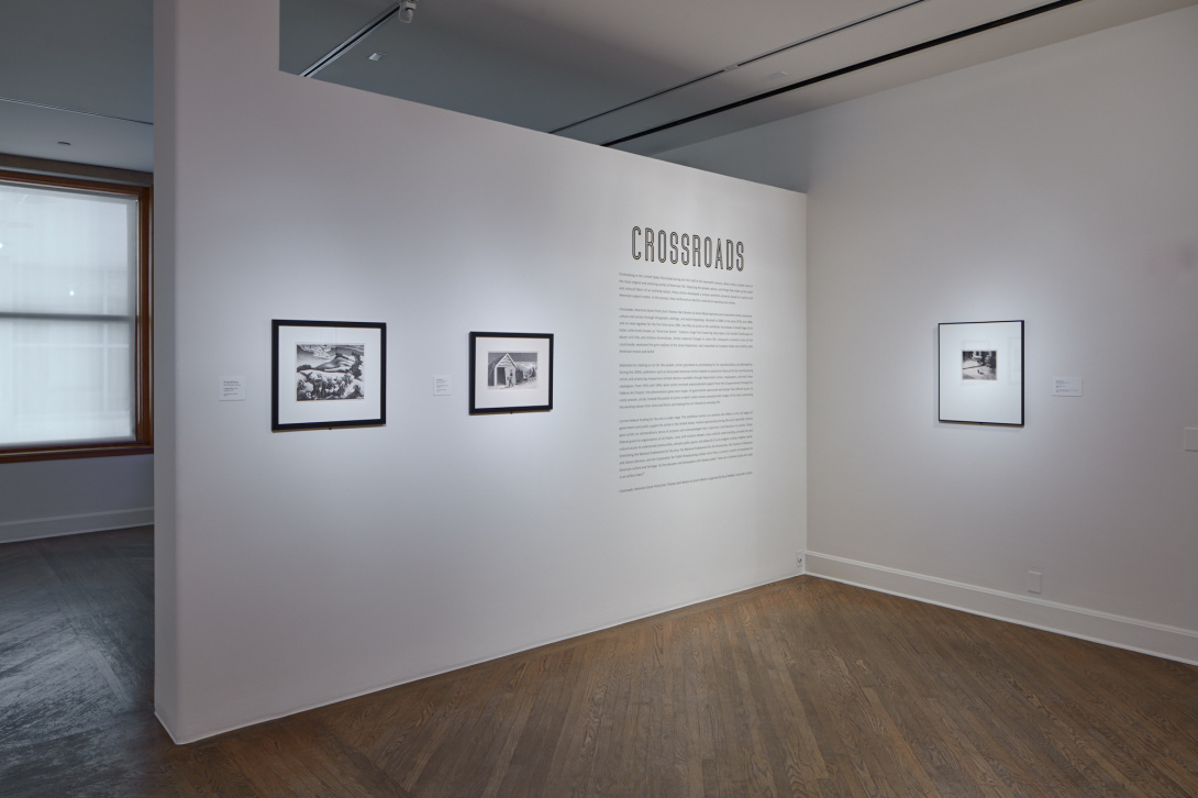 A picture showing a white wall outcrop of a room with three framed photographs and their descriptions. Also printed on the wall near the corner is the word "Crossroads" with a few paragraphs of text beneath.