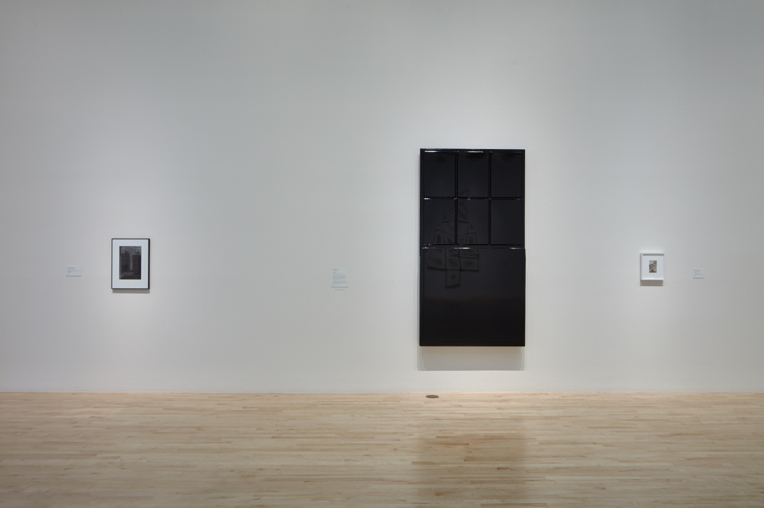 A large shiny black object that looks like a door hangs in the middle of a white gallery wall. To either side of it are small framed artworks, too small to decipher. The floor is made of light wood.