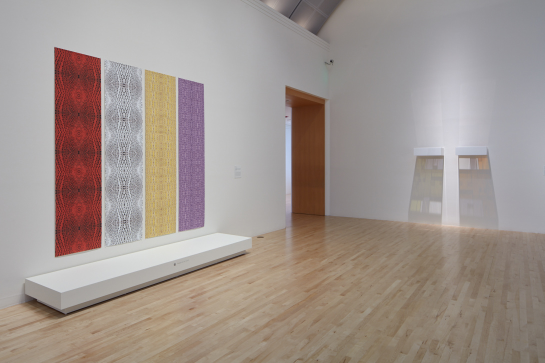 Four rectangular banners hang from a gallery wall. Each has a different color with a similar black pattern. Towards the back of the room are two white shelves. Light emanates from the shelves, casting shadows below. There is a door to another room.