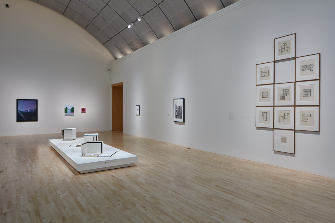 An open gallery space with three hanging art works on the back wall, three hanging on the right wall—one of which has a cluster of similar drawings togehter. A sculpture with various box-sized pieces sits on a white pedestal in the middle of the room.