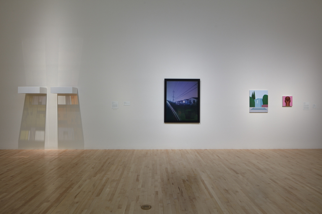 Four works of art hang in a gallery. To the left is a rectangular piece that has colors reflecting directly underneath it. Next to it is a photograph of a street at night. To the right are two paintings—a solid blue house and the back of a man's head with a pink background. 