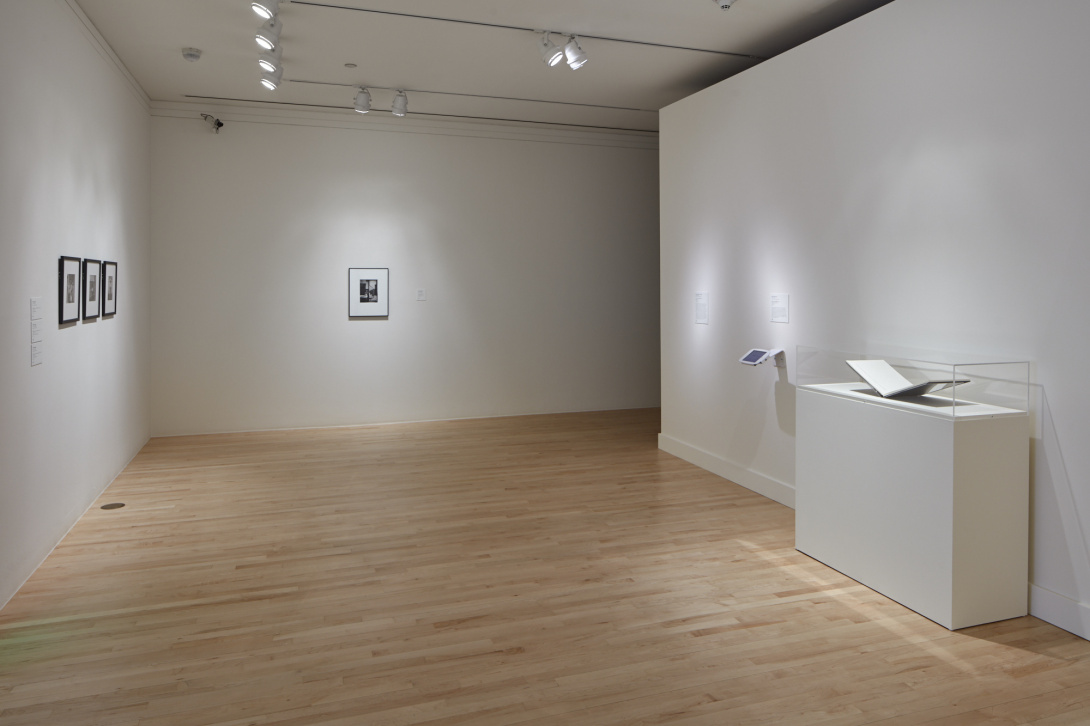 Open gallery space with three black and white photographs hanging on the left wall, another black and white photograph hanging on the back wall. On the right side there is a sculpture in a glass case that is shaped like an open book. 