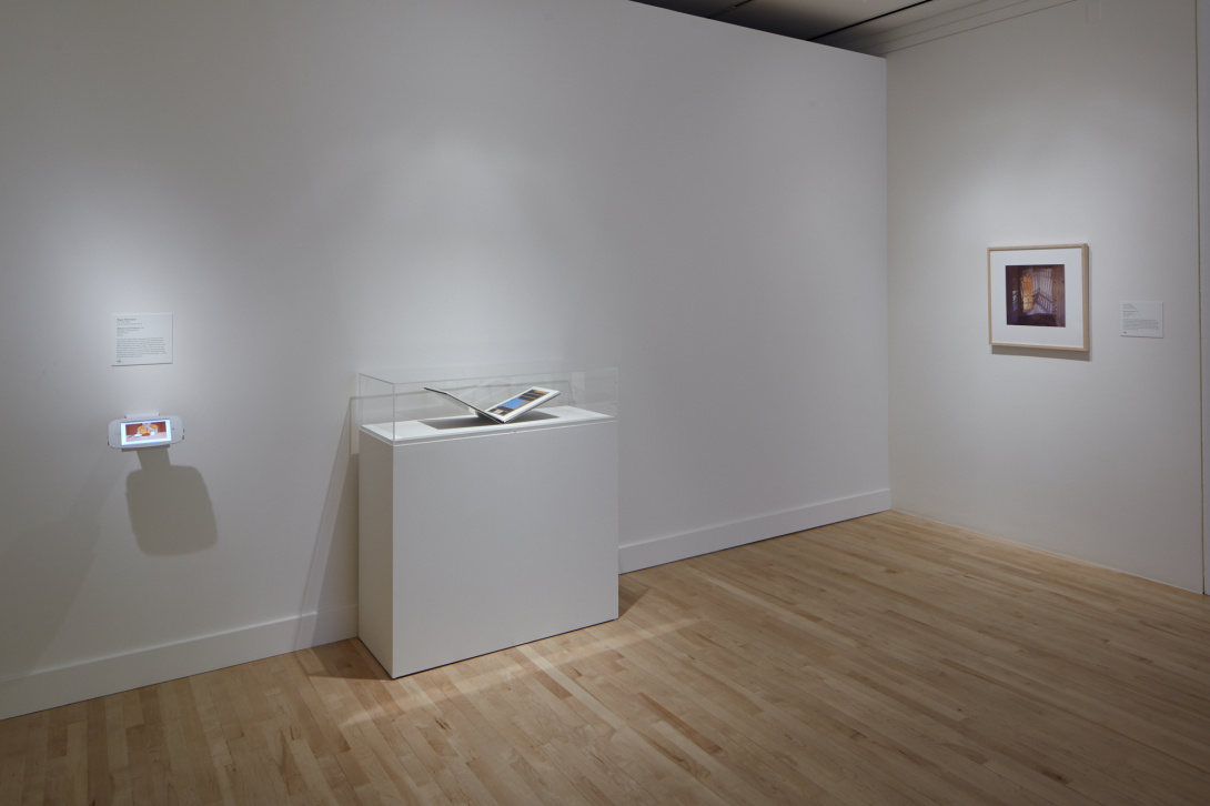 A pedestal with an open book on top of it that is protected by glass. To the left of the pedestal there is a tablet, and to the right there is a dark colored photograph hung on the wall. 