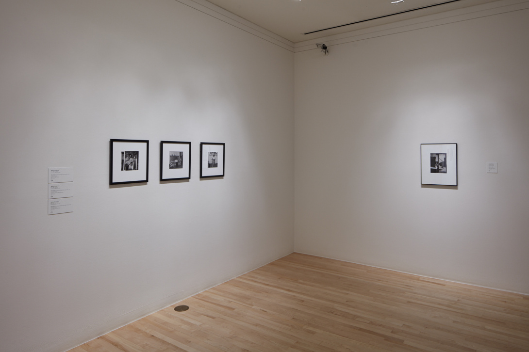 A corner of a gallery room with three framed artworks hung on the left side of the wall and on the right wall there is one framed artwork. 