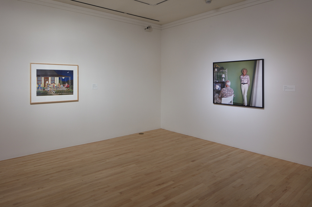 A photograph of the corner space of a gallery. A framed painting to hangs the left of the wall. To the right is a framed photograph.