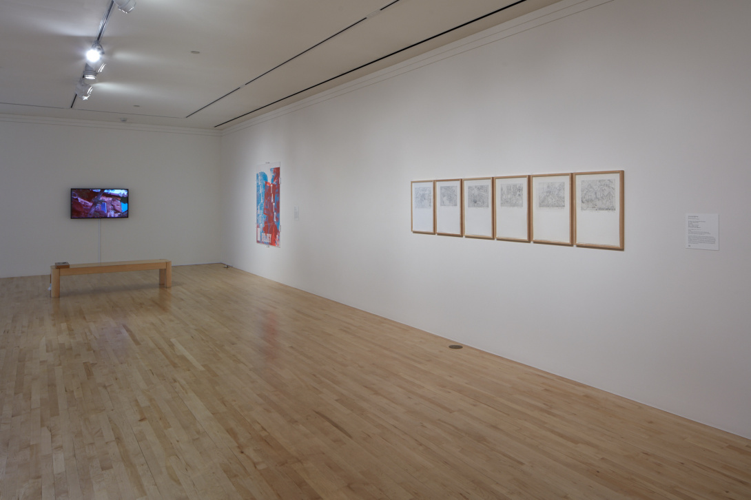 A photograph of a museum gallery. Towards the back is a lit television screen with a wooden bench in front of it. To the right side of the television, there is a large paper work and a set of six framed artworks. 