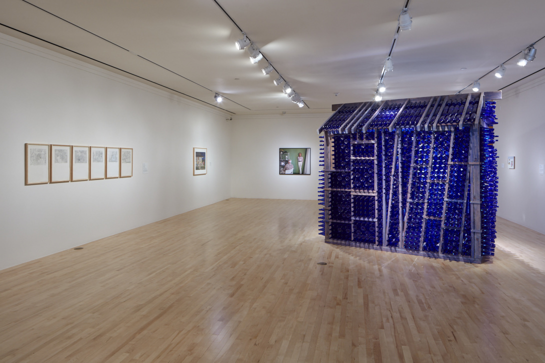 A photograph of a museum space. On view is a small-sized house made out of steel and blue glass bottles. There are nine works located behind this house. 
