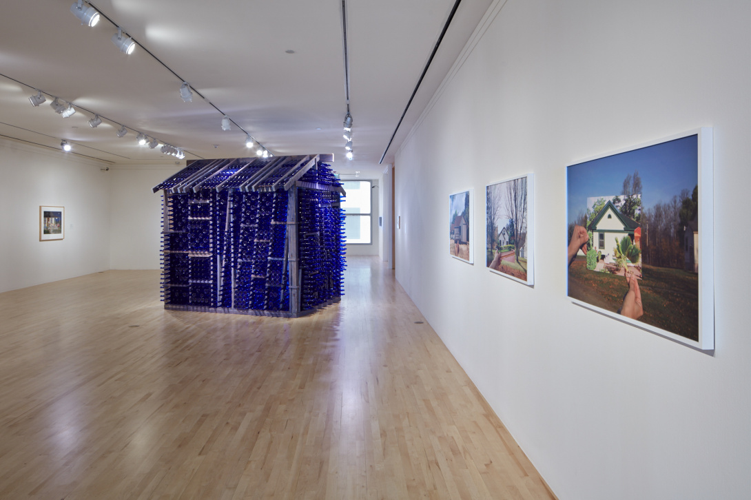 A photograph of a museum gallery. On view are three medium-sized photographic works of a house in the woodlands. A house sculpture made of steel and blue glass is located behind the photos.  
