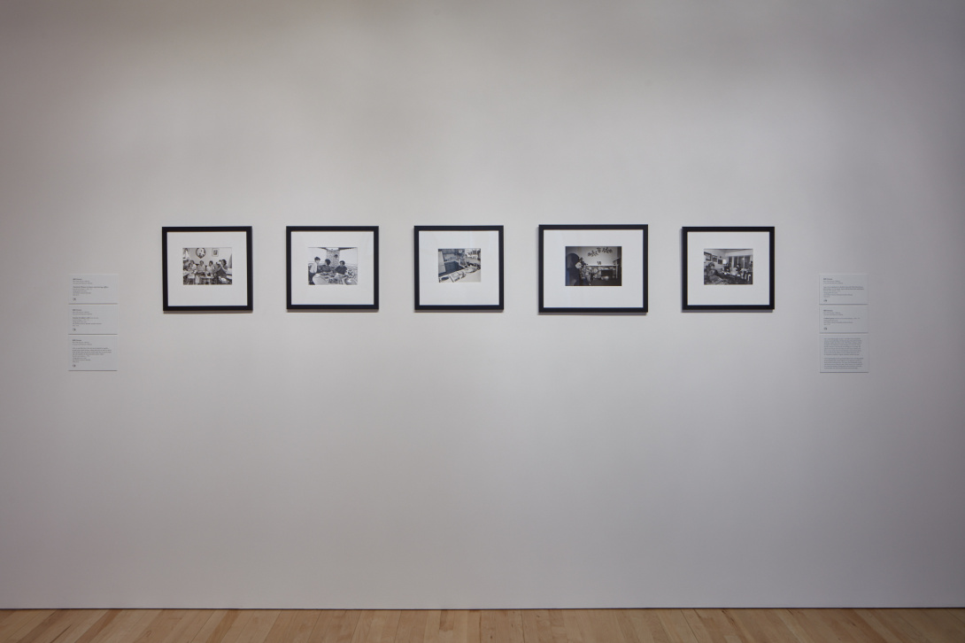 A photograph of a museum gallery space. On view are five black and white photographs mounted on a wall with black frames and information cards mounted on either side. They are in English, Spanish, and Vietnamese. 