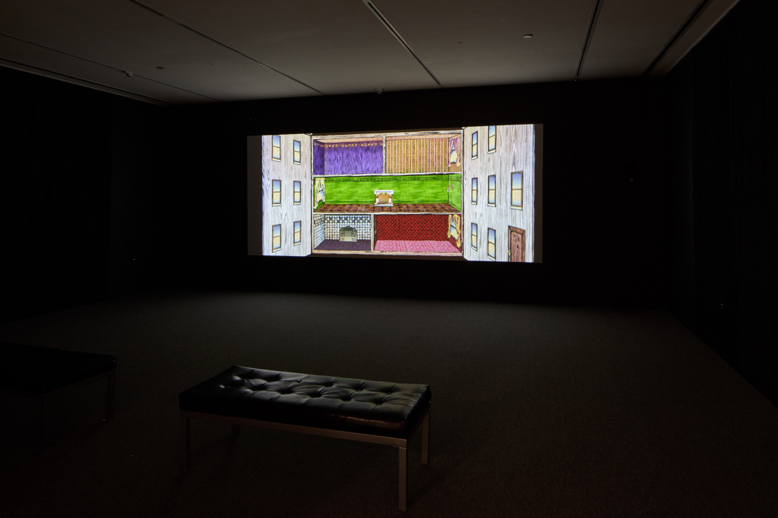 An open gallery space with the lights off, showing a projected image against the wall. There is a rectangular seat located in front of the screen.