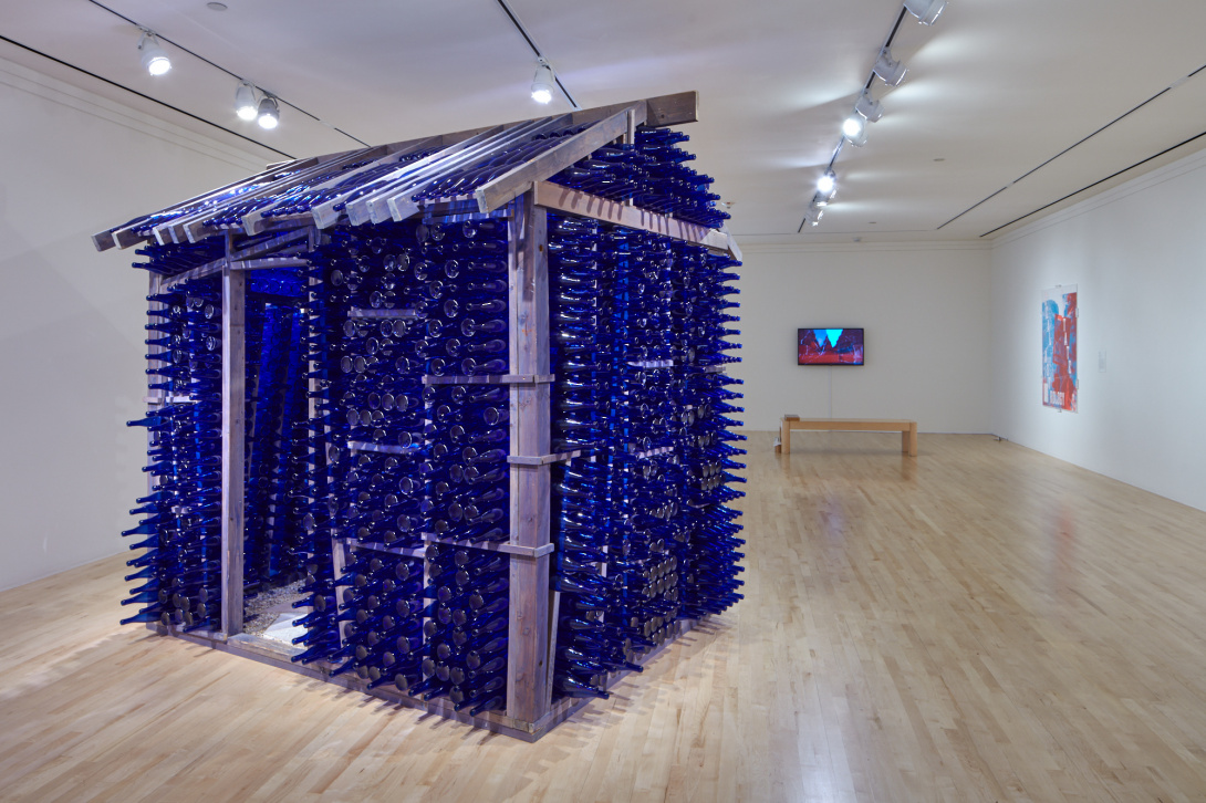 An open gallery space with a house-like figure made out of blue glass bottles and silver wood. In the background there are two art works mounted against the wall, one has a seat located in front of the video piece. 