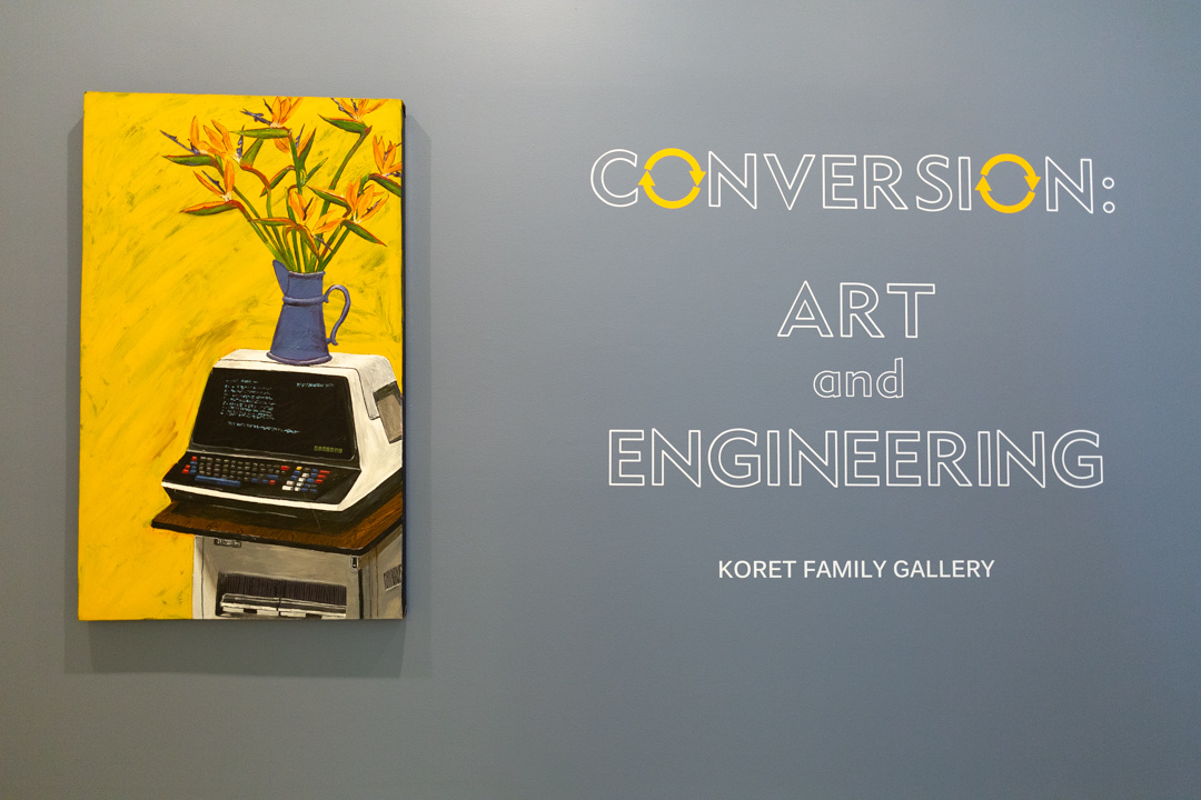 A painting with a bright yellow background features a watering can with flowers, sitting on top of an unidentifiable machine, which is on top of a copy machine. This painting hangs next to a title that reads "Conversion: Art and Engineering, Koret Family Gallery."