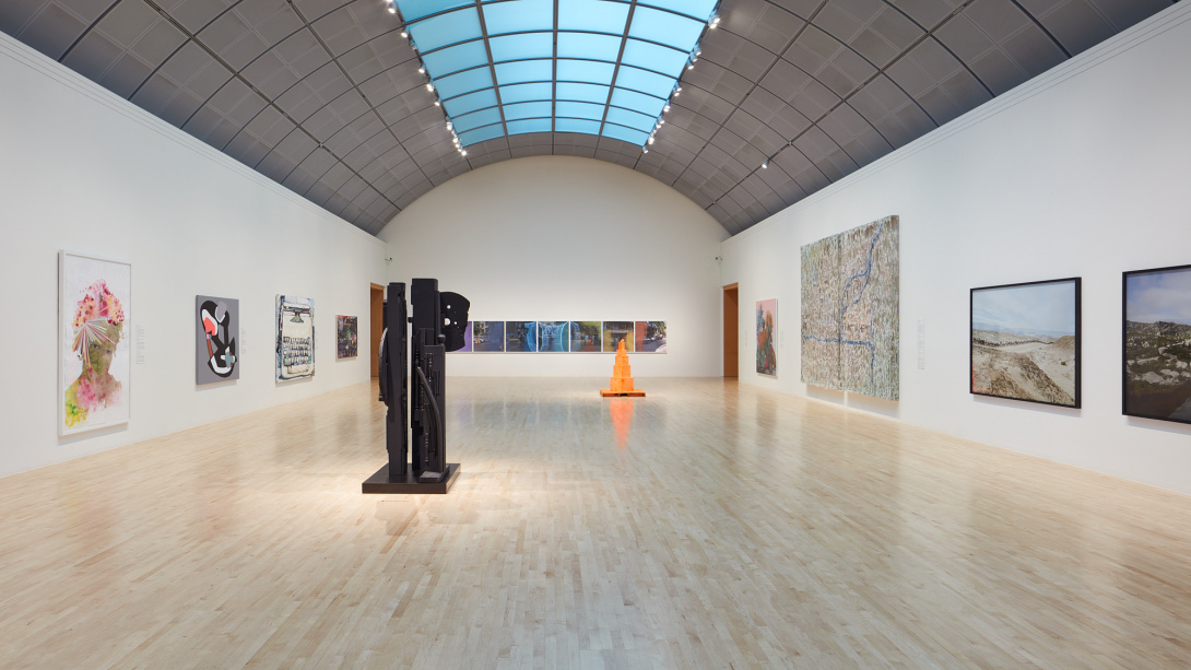 Museum gallery with tall curved ceiling with glass panels in middle. Two sculptures on the floor, one black and abstract forms and an orange minimal with boxes stacked. The left, far middle, and right walls have flat works of art.