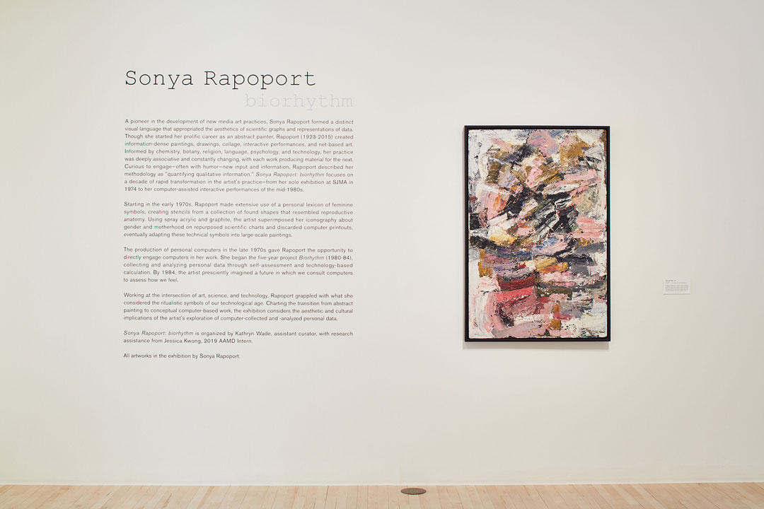 A gallery space with cream colored wall and a birch wood floor. On the left, the text reads, "Sonya Rapoport Biorhythm” with introductory texts of the exhibition. An abstract painting with pink, brown, black, and white is positioned on the right.