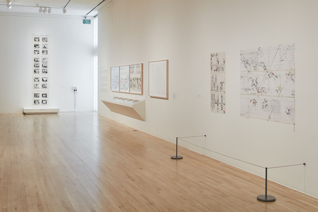 A gallery space with cream colored walls and a birch wood flooring. On the right side of the wall, there are several works hung on the wall and some in a display case. On the far back wall, there are 20 small black and white photographs displayed.