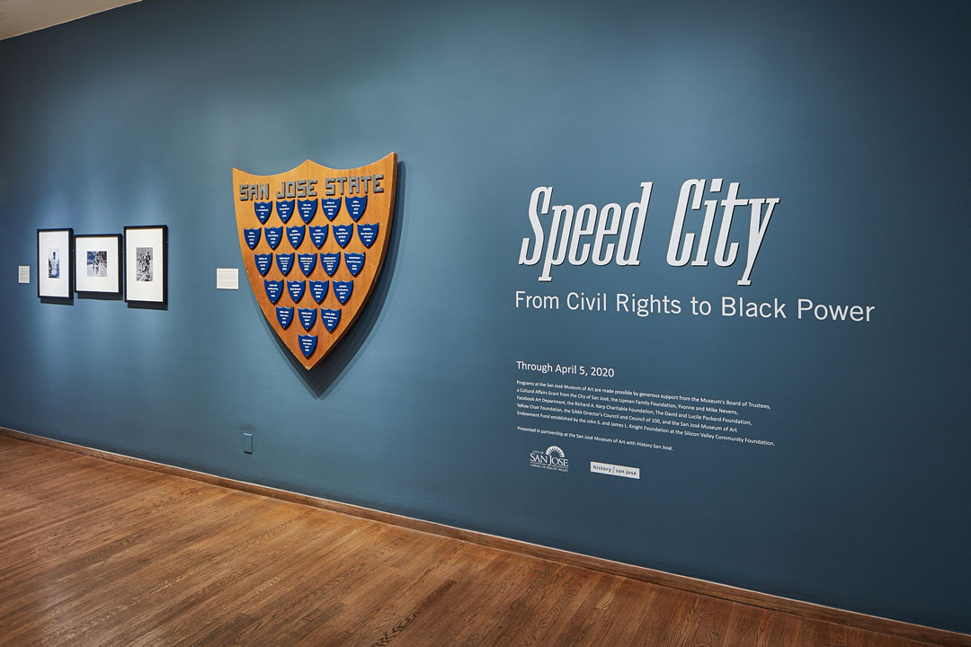 A large wooden badge shaped object filled with smaller blue badges hangs against a blue wall. To the right, "Speed City from Civil Rights to Black Power" reads in white vinyl. To the left are three black and white photographs—their subjects unclear.