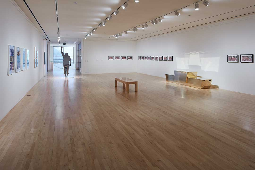 A gallery with a bench in the middle, artwork neatly placed on three walls and a sculpture of a man's silhouette standing at the back of the gallery with his arm raised high.