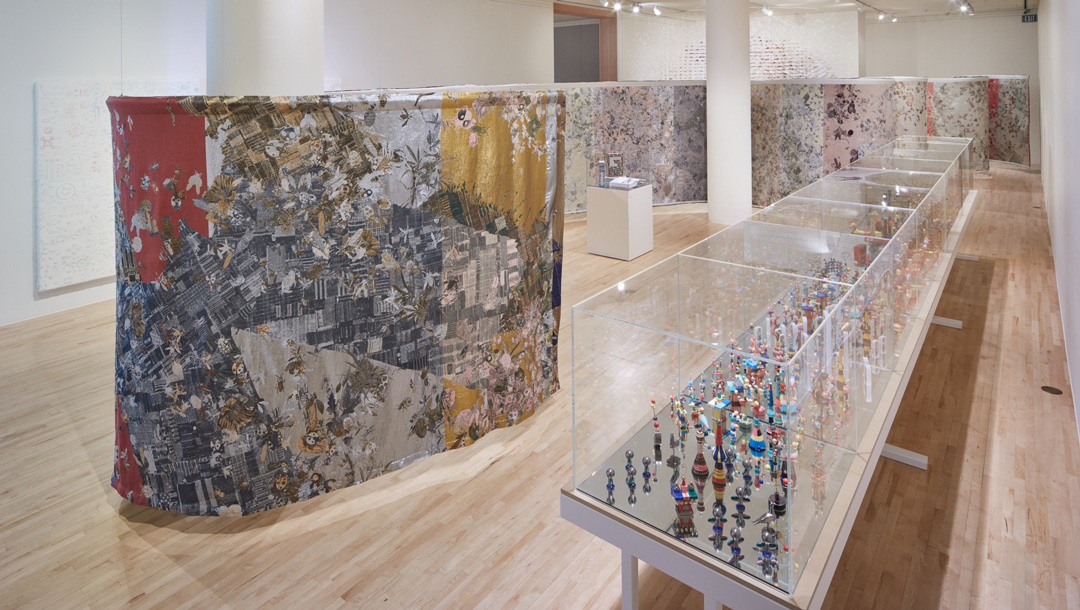 A long white cube gallery is bisected by a tall brightly colored and patterned tapestry that snakes through the space, preventing visibility from one side to the other. Adjacent to it, a long vitrine is filled with a gridded army of small bulbous statues.