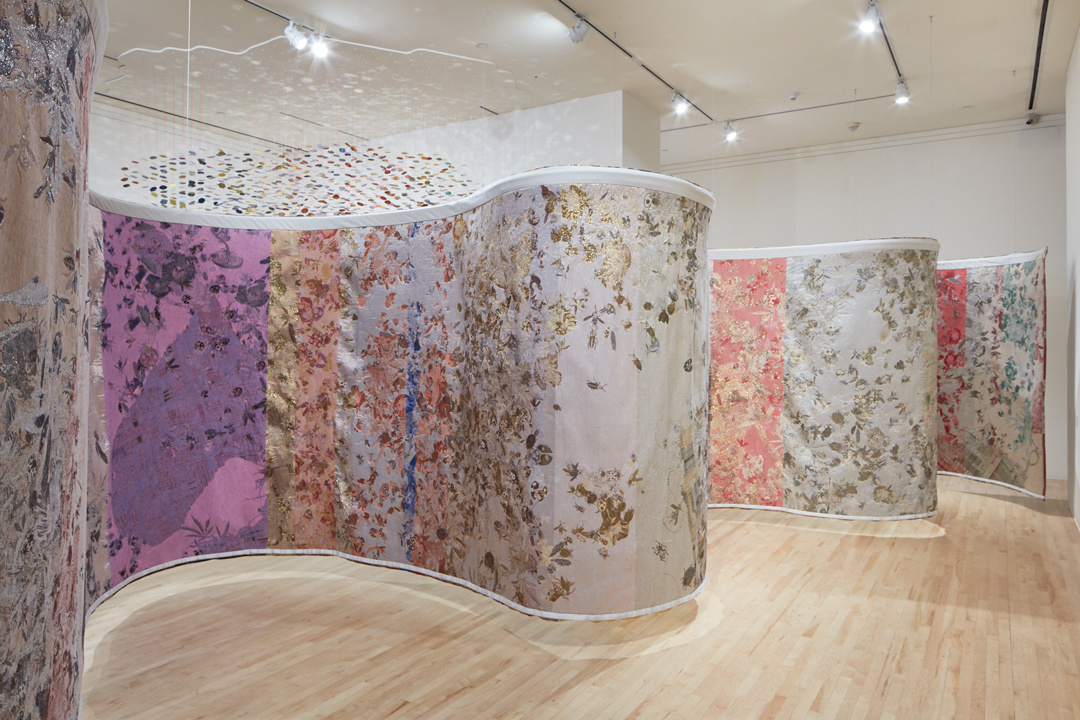 A brightly colored and abstractly patterned tapestry hangs from a gallery ceiling. It is so large that it barely fits in the space. Its serpentine shape divides the white room into three smaller nooks. Light passes underneath it and a confetti-like mobile is in the distance.