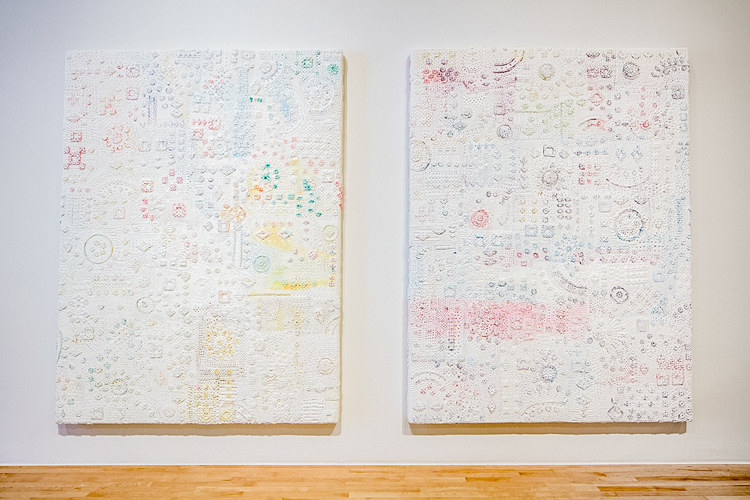Two nearly identical large monochrome white canvasses hang side-by-side on a gallery wall. Their textured surfaces are filled with shapes and curving lines. The texture is exaggerated by faint patches of pastel colors that become more saturated around the edges of some of the shapes.