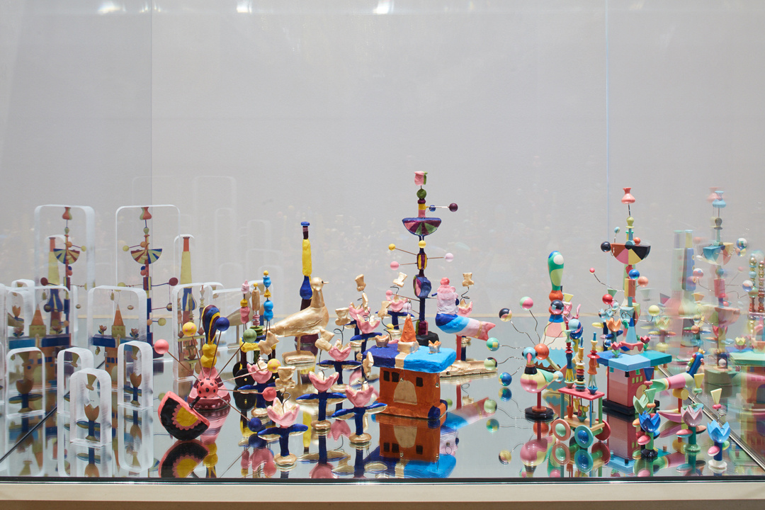 An array of whimsical brightly colored toy figurines are haphazardly arranged on a mirrored surface. Featuring colorful bobbles, gold painted birds, and rainbow fans, the toys range in size but none fills more than half the height of the glass box that contains them.