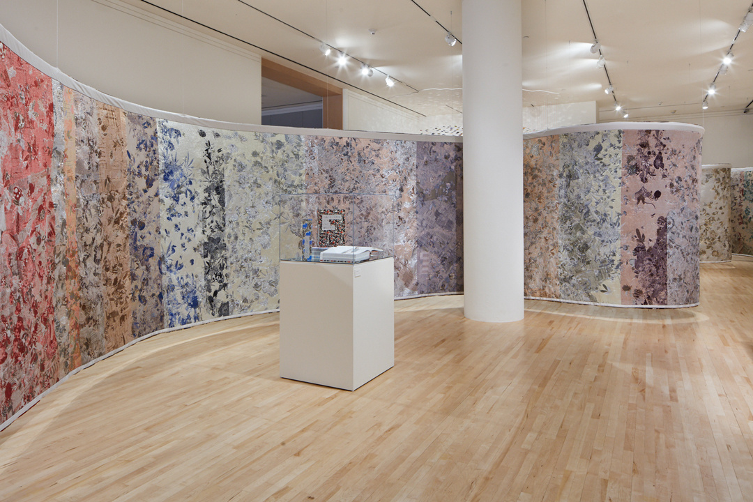 A large screen of abstractly patterned pastel colored panels curves through a white cube gallery and snakes around a white column at its center. In front of it, a square pedestal supports a glass vitrine containing three discrete objects.