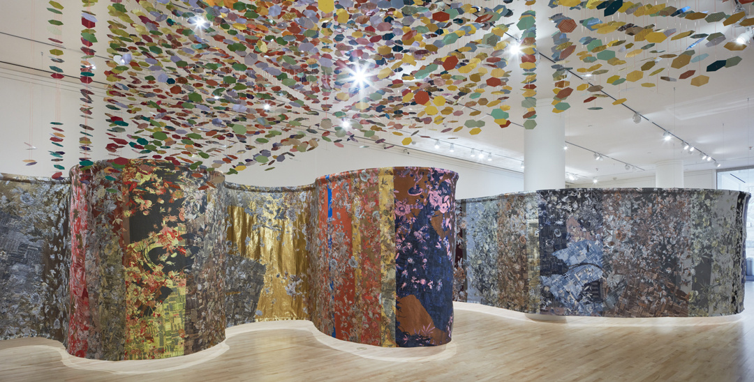 A large serpentine-shaped screen divides a gallery space into a series of curving nooks that invite close examination of its abstractly patterned bold colored panels. In the foreground, a mobile of scattered confetti-like disks matches its shimmering color scheme.