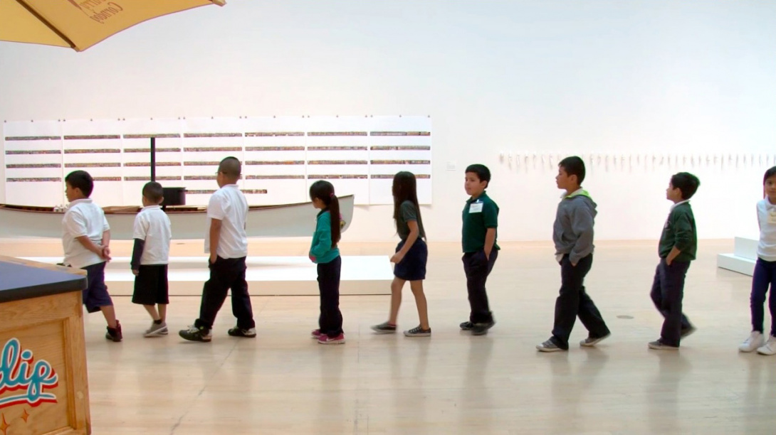Nine young children in school uniforms walk in single file with hands in their pockets across an art gallery. The children look at their surroundings and view the art on display including a sculpture of a large rowboat suspended mid-air. 