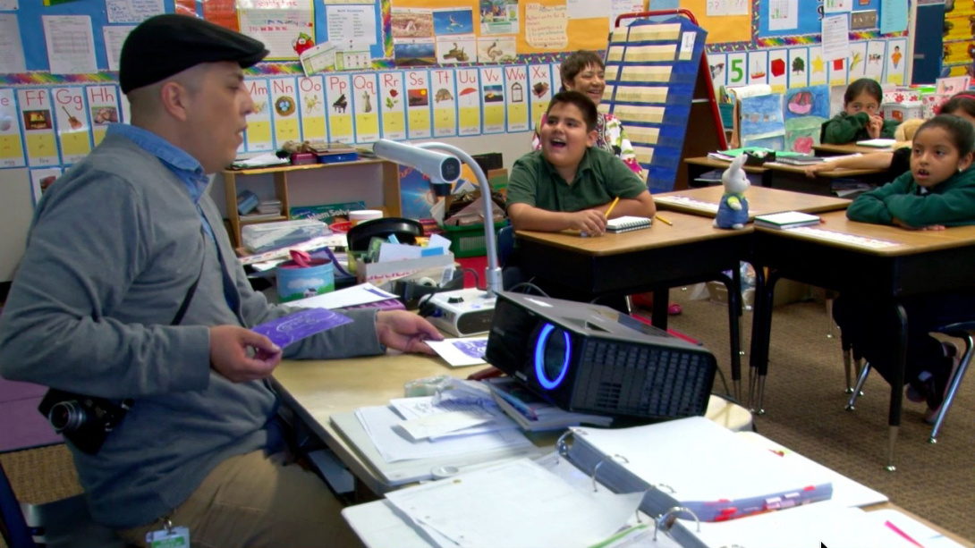 Four young children in school uniform sit at their desks while looking in awe and wonder at the front of the classroom. A seated adult  is wearing a hat, a camera across their chest, and holds photos while delivering a presentation with a projector. 