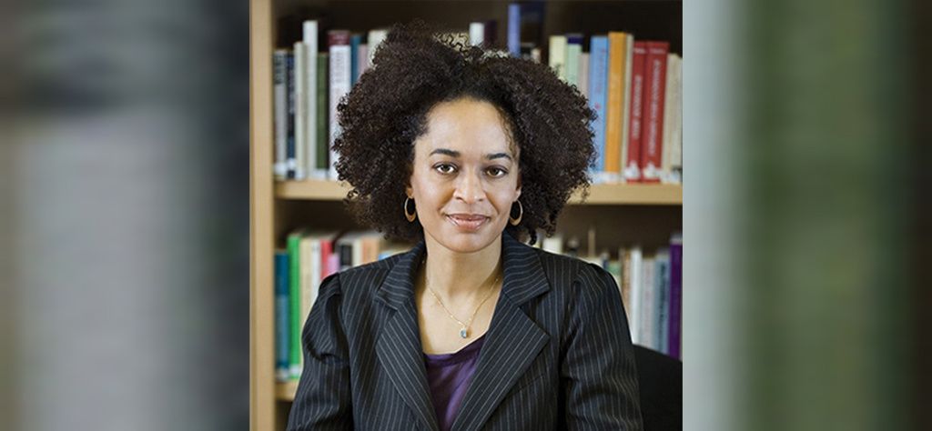 Woman with curly hair wearing a striped suit, sitting in front of a bookshelf.